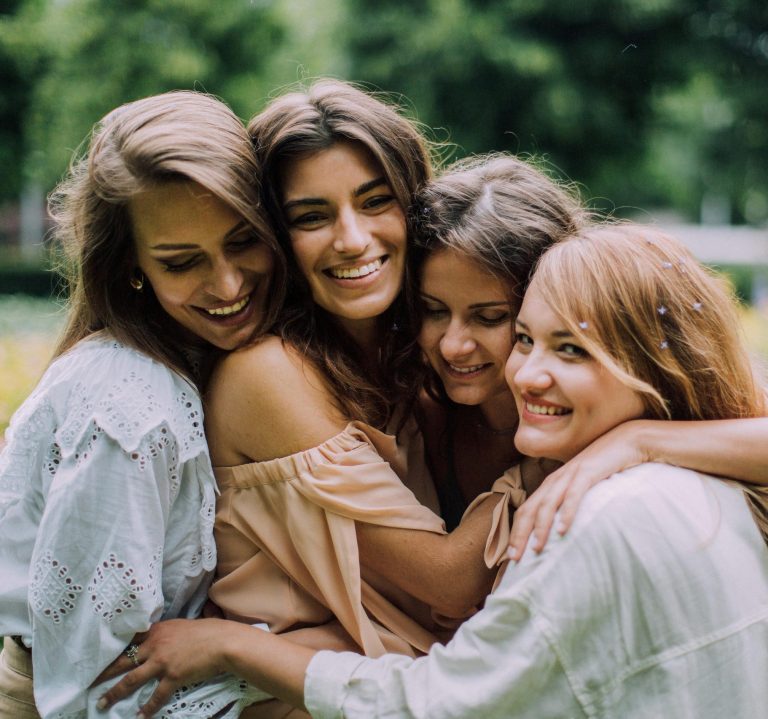 Four smiling women joyfully embrace in a lush green park during summer, showcasing genuine friendship and happiness.